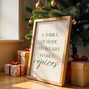 Puede incluir: Un letrero enmarcado en madera con el texto "A thrill of hope the weary world rejoices" en cursiva. El letrero está apoyado contra un árbol de Navidad con adornos dorados. Cajas de regalo con lazos rojos en primer plano.