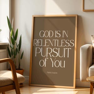 Può includere: Una stampa incorniciata con la scritta "GOD IS IN RELENTLESS PURSUIT of You" in bianco su sfondo marrone. La stampa è in una cornice di legno ed è appoggiata a un muro. La stampa è firmata "Patrick Keeron".