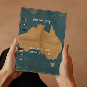 Pode incluir: Diário de viagem australiano com capa azul-petróleo apresentando um mapa da Austrália em castanho. O diário está aberto, revelando a encadernação em espiral. Os textos "jane and george" e "Aussie Travel Scrapbook Journal" são visíveis.