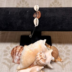 May include: A bracelet featuring three cowrie shells and a brown shell bead, strung on a silver-colored wire. The bracelet is displayed on a black velvet stand, with several seashells in the foreground.