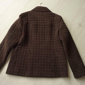 May include: A brown quilted jacket with a classic collar and long sleeves. The jacket has a square pattern throughout the fabric and is hanging on a metal hanger. The jacket is made of a soft, textured material.