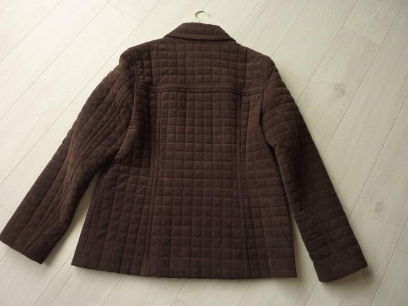 May include: A brown quilted jacket with a classic collar and long sleeves. The jacket has a square pattern throughout the fabric and is hanging on a metal hanger. The jacket is made of a soft, textured material.