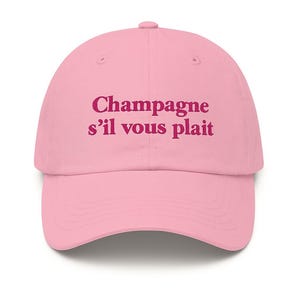 May include: A light pink baseball cap with the phrase "Champagne s'il vous plait" printed in a darker pink. The cap has a curved brim and a rounded crown, typical of a baseball cap.