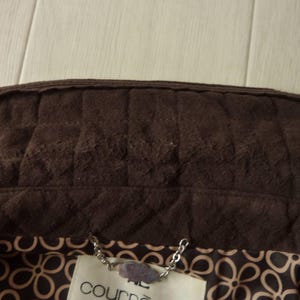 May include: Close-up of a brown suede jacket collar with quilted stitching. The interior lining features a floral pattern in shades of brown and beige. A silver chain and tag are visible at the bottom.