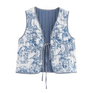 May include: Reversible quilted vest featuring a blue and white toile print with elephants, birds, and floral elements. The vest has a striped blue interior and a tie closure. The vest is sleeveless and has a V-neck.