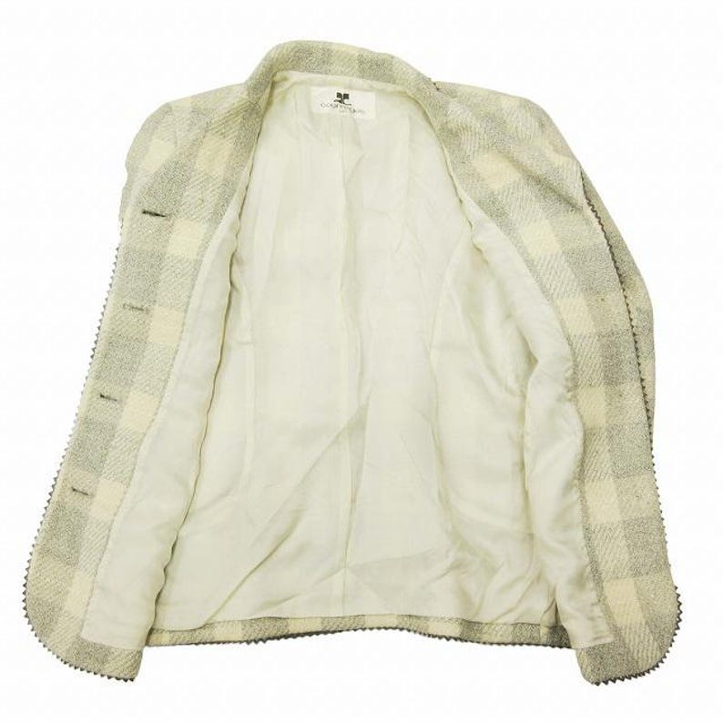 May include: A light-colored, open blazer with a checkered pattern in shades of cream and gray. The blazer has a notched collar, three buttons, and a decorative trim along the edges. The interior lining is a solid cream color.
