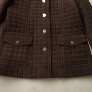 May include: A brown quilted jacket with a button-up front and two front pockets. The jacket has a diamond pattern and gold-colored buttons. The sleeves are long and the jacket appears to be made of a soft material.