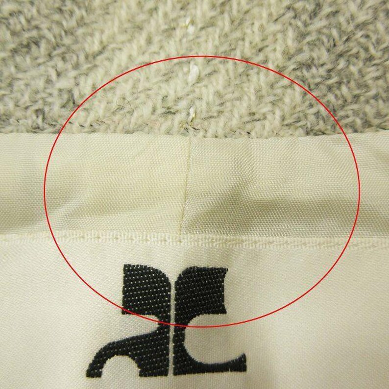May include: Close-up of a cream-colored fabric with a black logo at the bottom. The fabric has a textured appearance and a stitched seam. The logo is a stylized design.