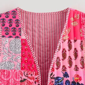 May include: A patchwork vest in shades of pink, featuring floral and paisley patterns. The vest has a V-neck and a tie closure. The design includes vertical stripes and quilted sections, creating a vibrant and textured look.