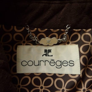 May include: Close-up of a brown jacket interior with a floral pattern lining. A white label with the brand name "courr&egrave;ges" in black lettering is visible. A silver chain and a decorative clasp are also present.