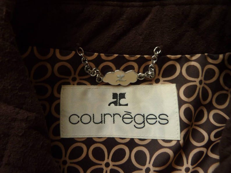 May include: Close-up of a brown jacket interior with a floral pattern lining. A white label with the brand name "courr&egrave;ges" in black lettering is visible. A silver chain and a decorative clasp are also present.