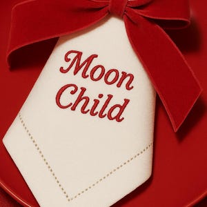 Puede incluir: Una servilleta de lino blanco doblada con las palabras "Moon Child" bordadas en rojo, acentuada por un lazo de terciopelo rojo. La servilleta descansa sobre un plato rojo, creando una presentación festiva y elegante.