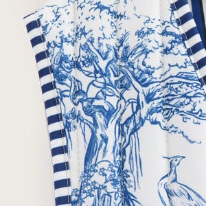 May include: Close-up of a white fabric with a blue toile print featuring a tree, bird, and floral design. The edges are trimmed with blue and white striped fabric. The overall aesthetic is classic and elegant.
