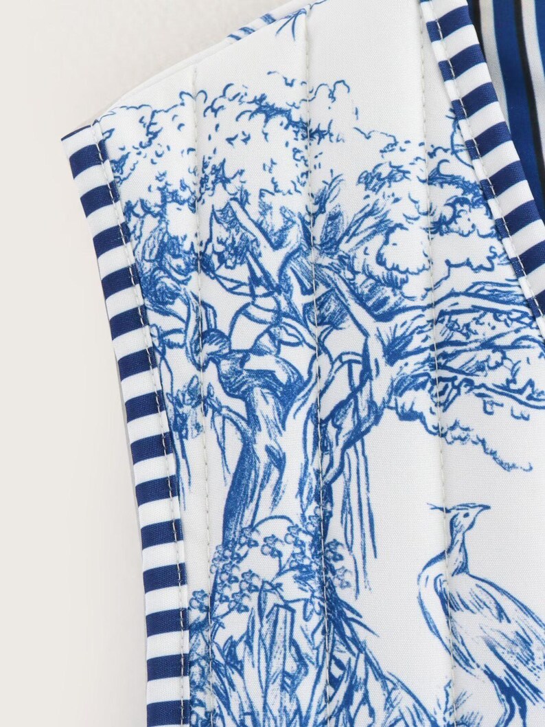 May include: Close-up of a white fabric with a blue toile print featuring a tree, bird, and floral design. The edges are trimmed with blue and white striped fabric. The overall aesthetic is classic and elegant.