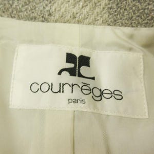 May include: Close-up of a white label with the black Courr&egrave;ges logo and text "courr&egrave;ges paris". The label is sewn into a light-colored fabric, possibly a jacket or coat lining. The background is a textured, light gray material.
