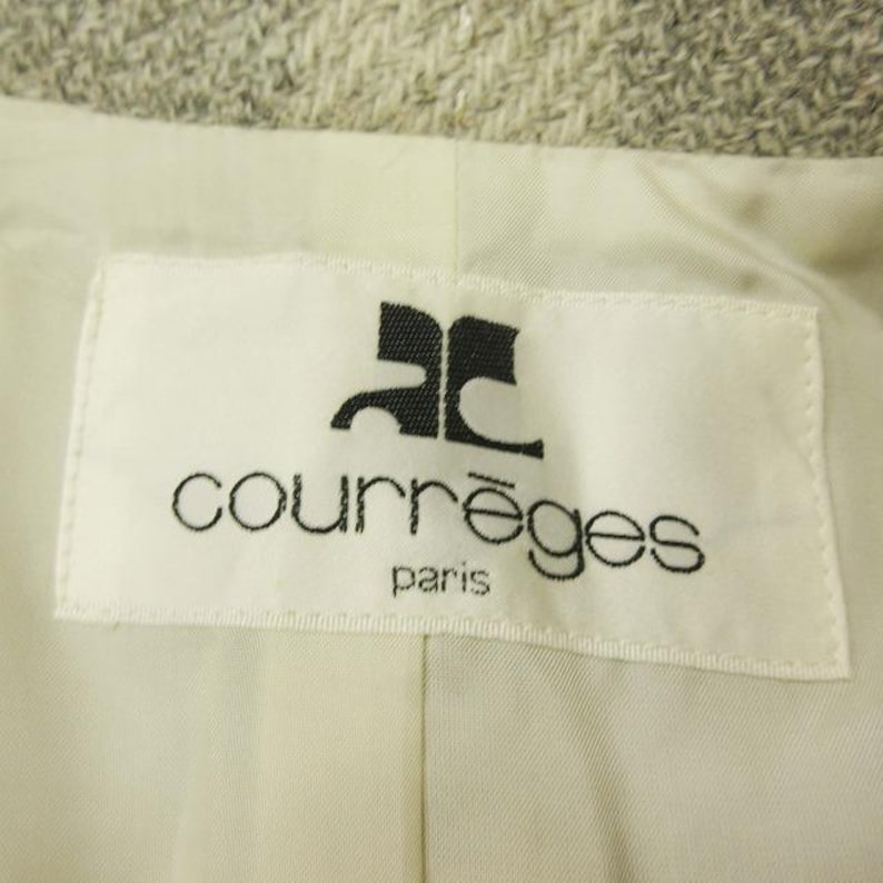 May include: Close-up of a white label with the black Courr&egrave;ges logo and text "courr&egrave;ges paris". The label is sewn into a light-colored fabric, possibly a jacket or coat lining. The background is a textured, light gray material.