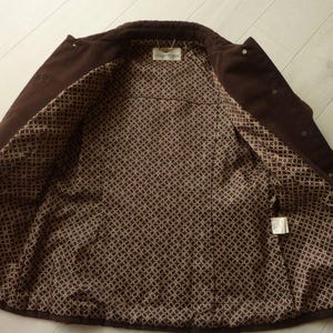 May include: A dark brown corduroy jacket with a patterned interior lining. The jacket is open, revealing the lining with a repeating circular design in shades of brown and tan. The jacket has a collar and button closures.