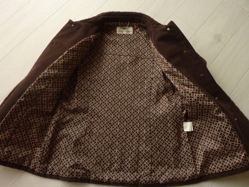 May include: A dark brown corduroy jacket with a patterned interior lining. The jacket is open, revealing the lining with a repeating circular design in shades of brown and tan. The jacket has a collar and button closures.