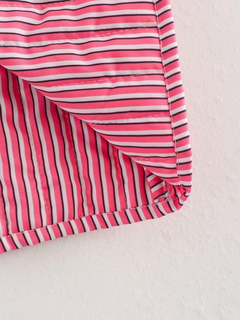 May include: Close-up of a section of fabric with a pink, black, and white striped pattern. The fabric is folded over, revealing the interior lining. The stripes are vertical and evenly spaced, with a stitched seam visible.