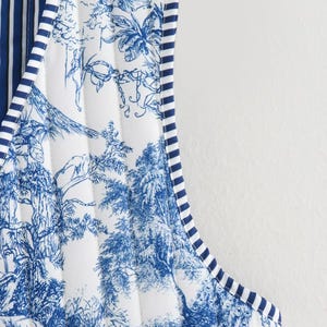 May include: Close-up of a quilted vest featuring a blue and white toile pattern depicting a landscape scene. The vest has a striped trim in navy blue and white. The interior lining has vertical navy blue and white stripes.