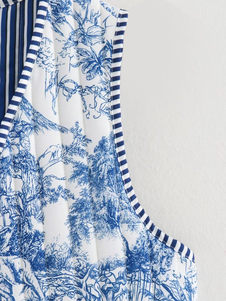 May include: Close-up of a quilted vest featuring a blue and white toile pattern depicting a landscape scene. The vest has a striped trim in navy blue and white. The interior lining has vertical navy blue and white stripes.