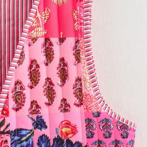 May include: A sleeveless, quilted garment featuring a patchwork design. The garment showcases a variety of pink hues, with floral, paisley, and striped patterns. The edges are trimmed with a striped border, creating a vibrant and detailed aesthetic.
