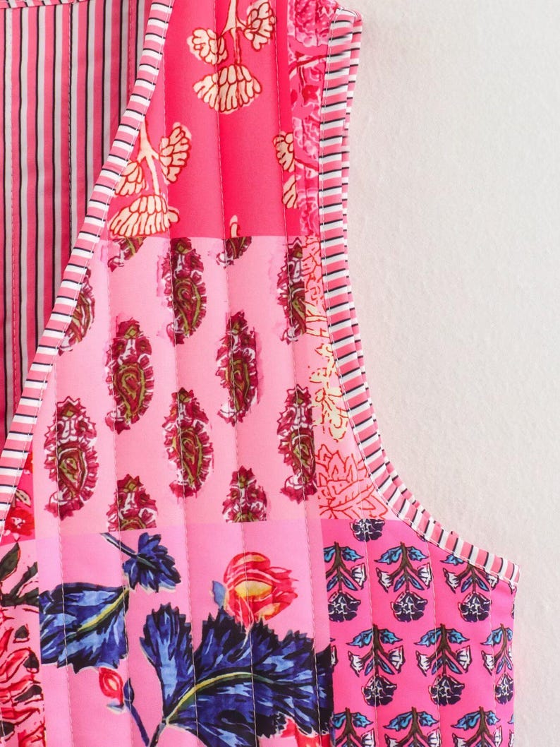 May include: A sleeveless, quilted garment featuring a patchwork design. The garment showcases a variety of pink hues, with floral, paisley, and striped patterns. The edges are trimmed with a striped border, creating a vibrant and detailed aesthetic.
