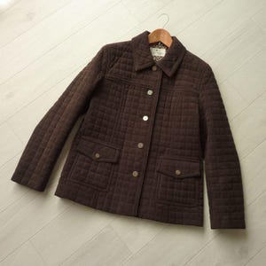 May include: A brown quilted jacket with a classic design. The jacket features a button-up front, two front pockets, and a collared neckline. The jacket is hanging on a wooden hanger.