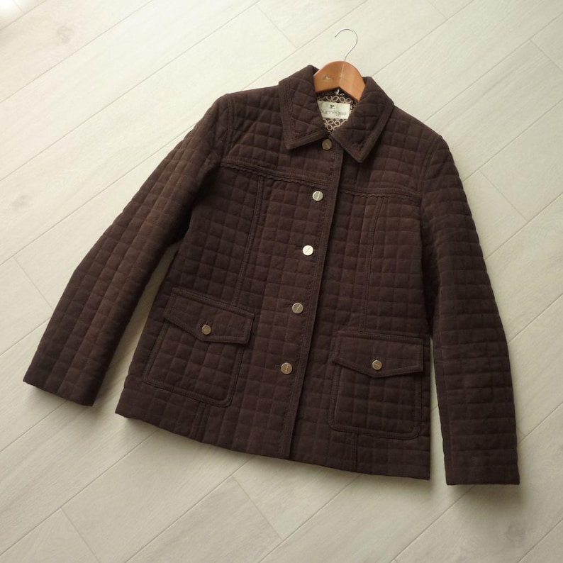 May include: A brown quilted jacket with a classic design. The jacket features a button-up front, two front pockets, and a collared neckline. The jacket is hanging on a wooden hanger.