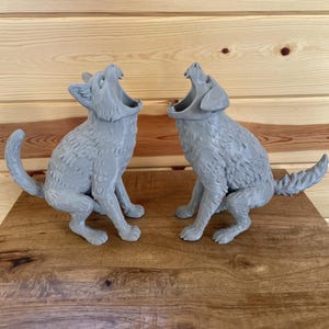 May include: Two light grey, three-dimensional figures of animals in a seated, howling position with mouths wide open. The figures feature textured fur details, pointed ears, and tails extending outward, crafted with a matte finish surface.
