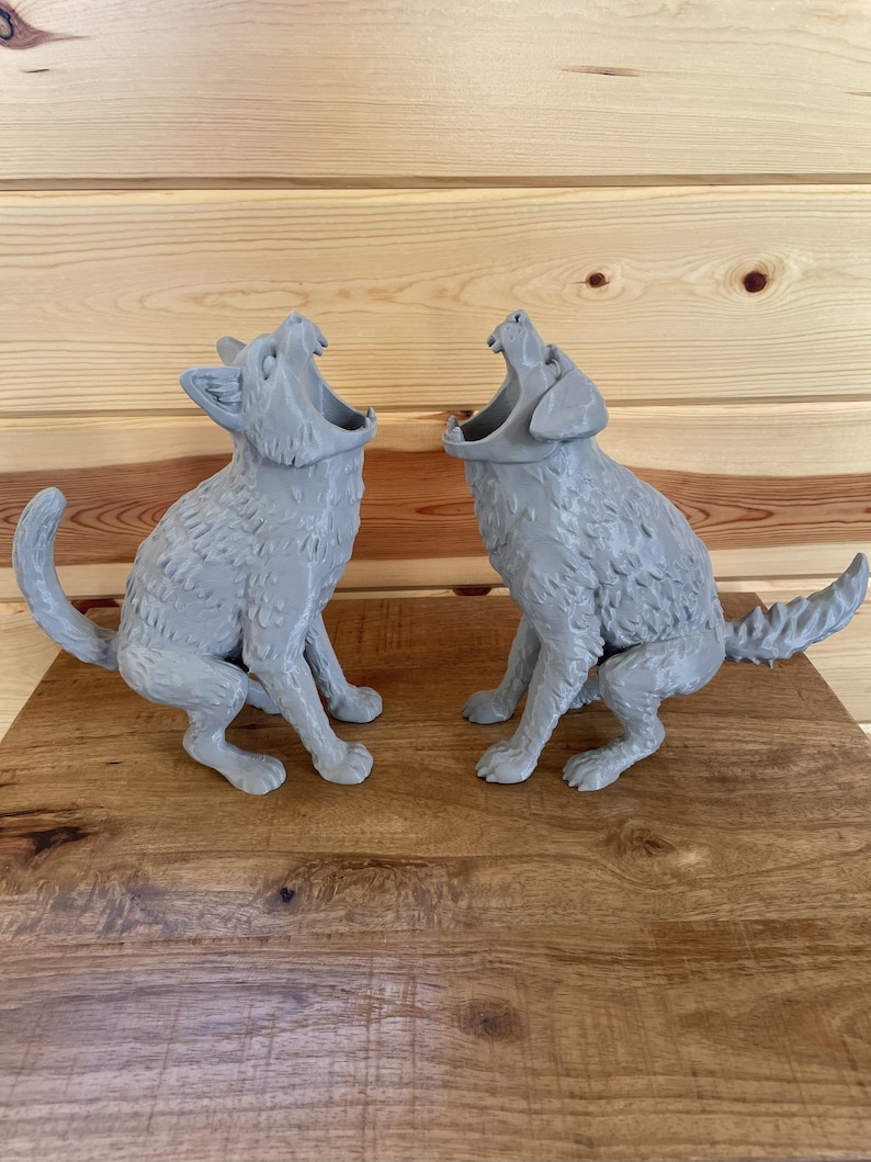 May include: Two light grey, three-dimensional figures of animals in a seated, howling position with mouths wide open. The figures feature textured fur details, pointed ears, and tails extending outward, crafted with a matte finish surface.