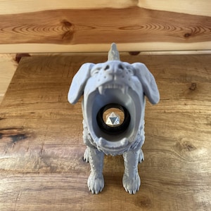 May include: A light grey 3D printed figurine of a dog with its mouth wide open, revealing a hollow interior that holds a single silver twenty-sided die. The dog has textured fur, pointed ears, and a small horn-like protrusion on its head.