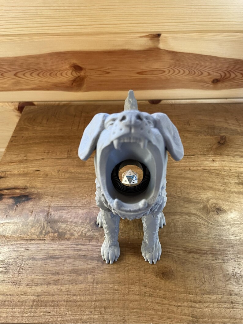 May include: A light grey 3D printed figurine of a dog with its mouth wide open, revealing a hollow interior that holds a single silver twenty-sided die. The dog has textured fur, pointed ears, and a small horn-like protrusion on its head.