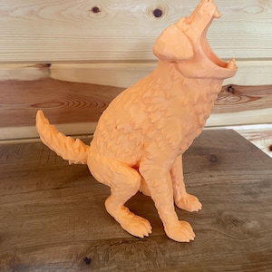May include: A light orange, three-dimensional figurine of a dog in a seated position with its head tilted upwards and mouth wide open. The sculpture features a textured, faceted surface that mimics fur, with a pointed tail and visible paws.