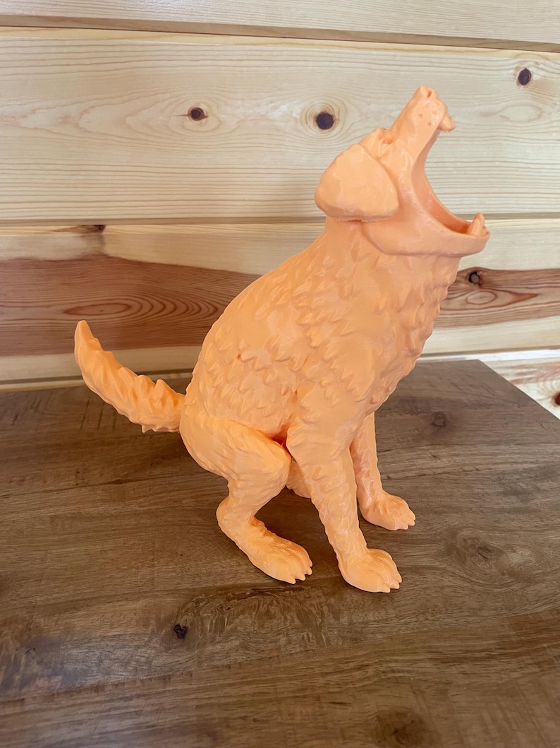 May include: A light orange, three-dimensional figurine of a dog in a seated position with its head tilted upwards and mouth wide open. The sculpture features a textured, faceted surface that mimics fur, with a pointed tail and visible paws.