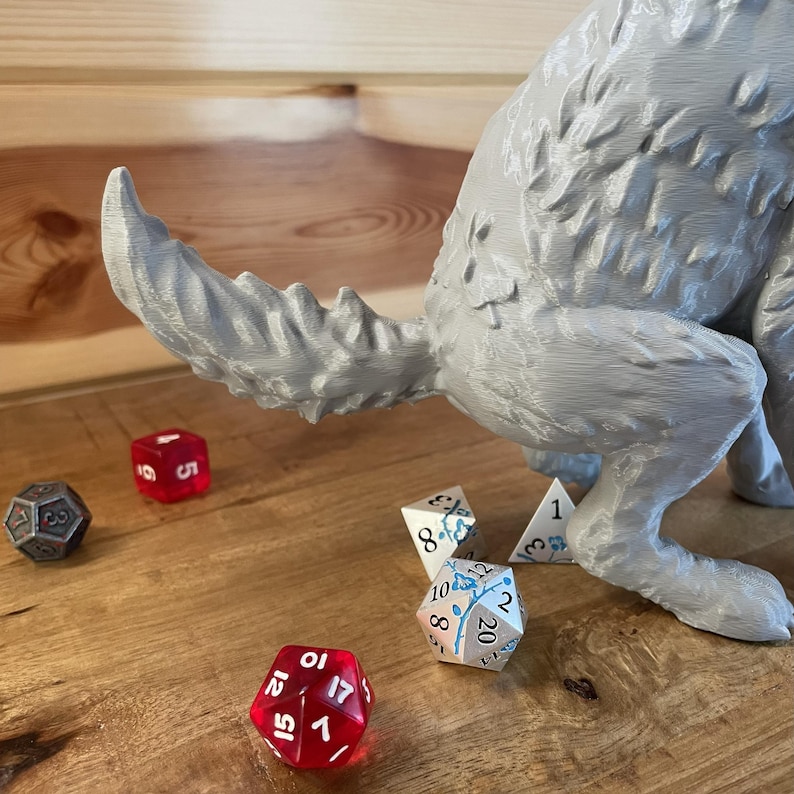 May include: A grey 3D-printed wolf figurine with a textured fur pattern, positioned with its tail extended. Several multi-sided gaming dice are scattered nearby, including a translucent red twenty-sided die and a white die with blue floral details.