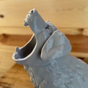 May include: A 3D printed grey figurine of a howling wolf with its head tilted upwards and mouth wide open. The surface of the object displays visible horizontal layer lines characteristic of the additive manufacturing process, showing the snout, ears, and fur texture.