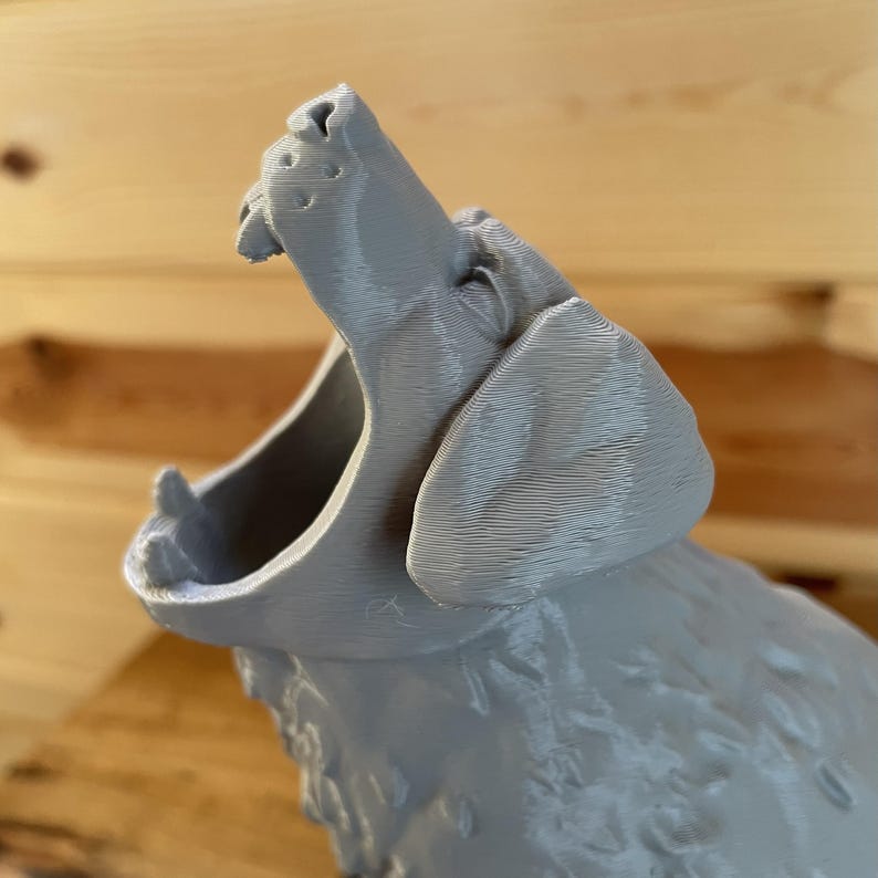 May include: A 3D printed grey figurine of a howling wolf with its head tilted upwards and mouth wide open. The surface of the object displays visible horizontal layer lines characteristic of the additive manufacturing process, showing the snout, ears, and fur texture.