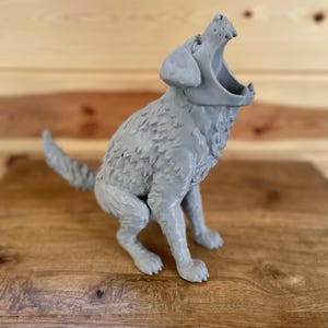 May include: A grey 3D printed figurine of a dog in a seated position with its head tilted upwards and mouth wide open. The dog has textured fur detailing across its body, a pointed tail, and visible teeth within the open jaw, rendered in a solid matte grey finish.