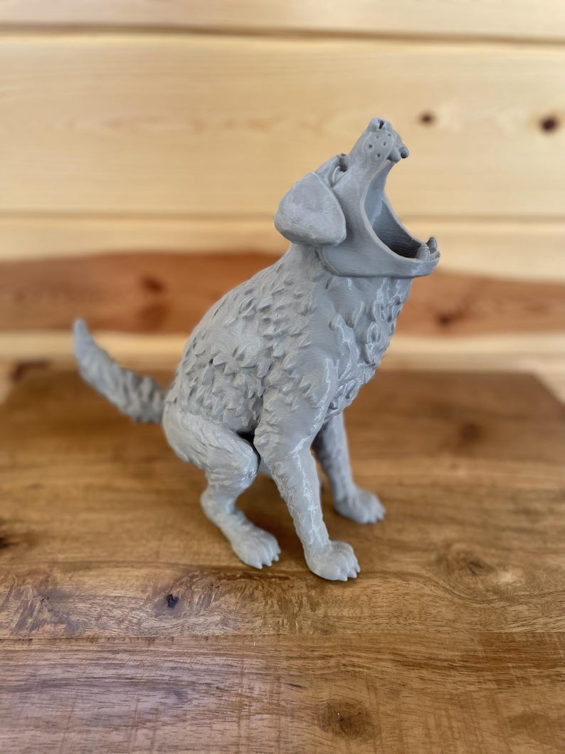 May include: A grey 3D printed figurine of a dog in a seated position with its head tilted upwards and mouth wide open. The dog has textured fur detailing across its body, a pointed tail, and visible teeth within the open jaw, rendered in a solid matte grey finish.