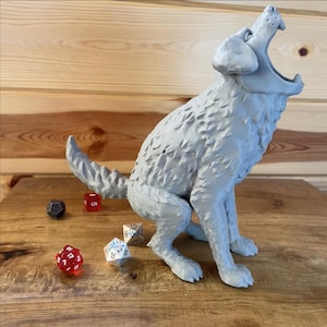 May include: A grey, 3D-printed wolf figurine in a howling pose. The wolf is sitting with its mouth open, and its tail is curled. Several polyhedral dice in red, white, and black are scattered around the base of the figurine. The figurine is on a wooden surface.