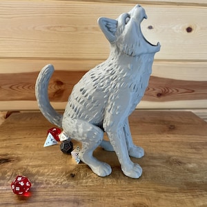 May include: A light gray 3D-printed cat figurine in a seated position with its head tilted back and mouth wide open. The cat has textured fur detailing and is surrounded by several multi-sided polyhedral dice in red, white, and black colors.