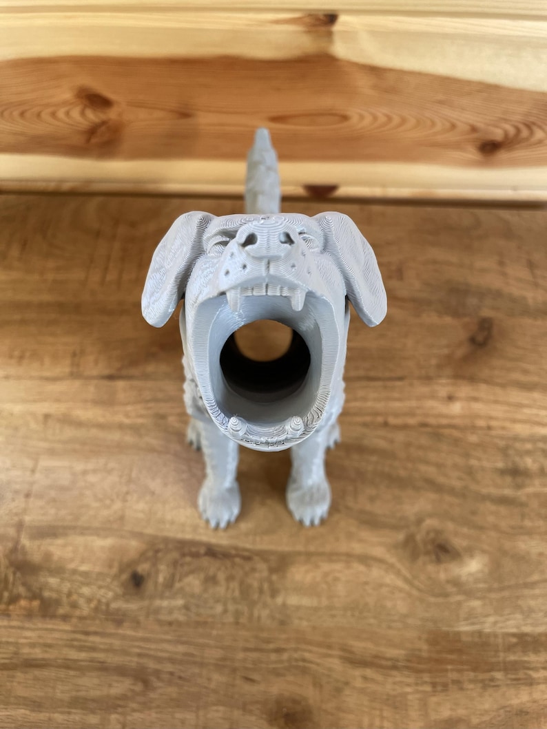 May include: A light grey 3D printed dog figurine with a hollow, open mouth design. The canine figure features visible horizontal layer lines from the printing process, floppy ears, and a textured surface, standing in a forward-facing position.