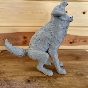 May include: A grey 3D printed figurine of a wolf in a seated position with its head tilted upwards and mouth wide open as if howling. The surface of the wolf displays a textured, layered finish characteristic of additive manufacturing processes.