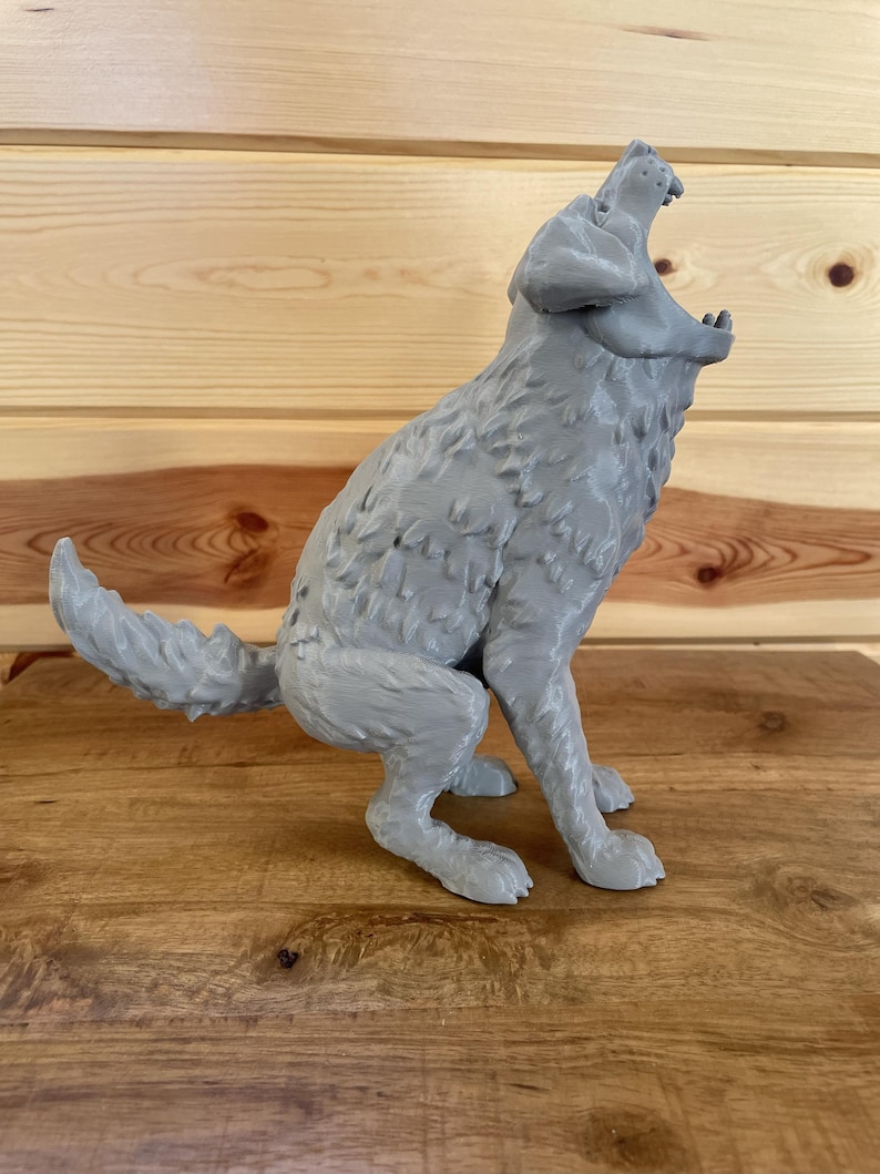 May include: A grey 3D printed figurine of a wolf in a seated position with its head tilted upwards and mouth wide open as if howling. The surface of the wolf displays a textured, layered finish characteristic of additive manufacturing processes.
