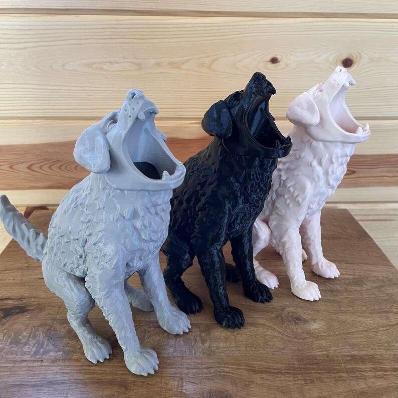 May include: Three dog-shaped containers in light grey, black, and off-white. Each figure is depicted in a seated position with its head tilted upward and mouth wide open, revealing a hollow interior. The textured surfaces show detailed fur.