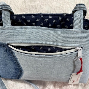 May include: A denim tote bag with a light blue exterior and a dark blue floral patterned interior. The bag features a front zipper pocket with a white zipper and a red tag. The bag has two handles and a shoulder strap.