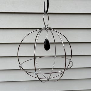 Puede incluir: Una escultura colgante esférica de metal plateado con una piedra negra de forma irregular suspendida en el centro. La escultura está compuesta por varillas metálicas circulares que se cruzan, creando una estructura similar a una jaula. Cuelga de un gancho negro.