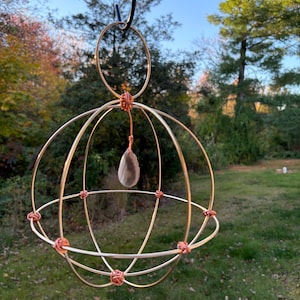 May include: A handcrafted, spherical hanging sculpture made of gold-colored metal wire. The wire is intricately woven and knotted with copper-colored wire. A teardrop-shaped, translucent stone hangs from the center. The sculpture is suspended from a black hook.