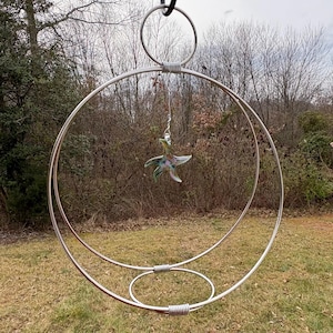 May include: A large, silver metal outdoor sculpture featuring a series of concentric circles and a hanging, colorful glass starfish. The sculpture is suspended from a black cable and set against a backdrop of trees and grass.